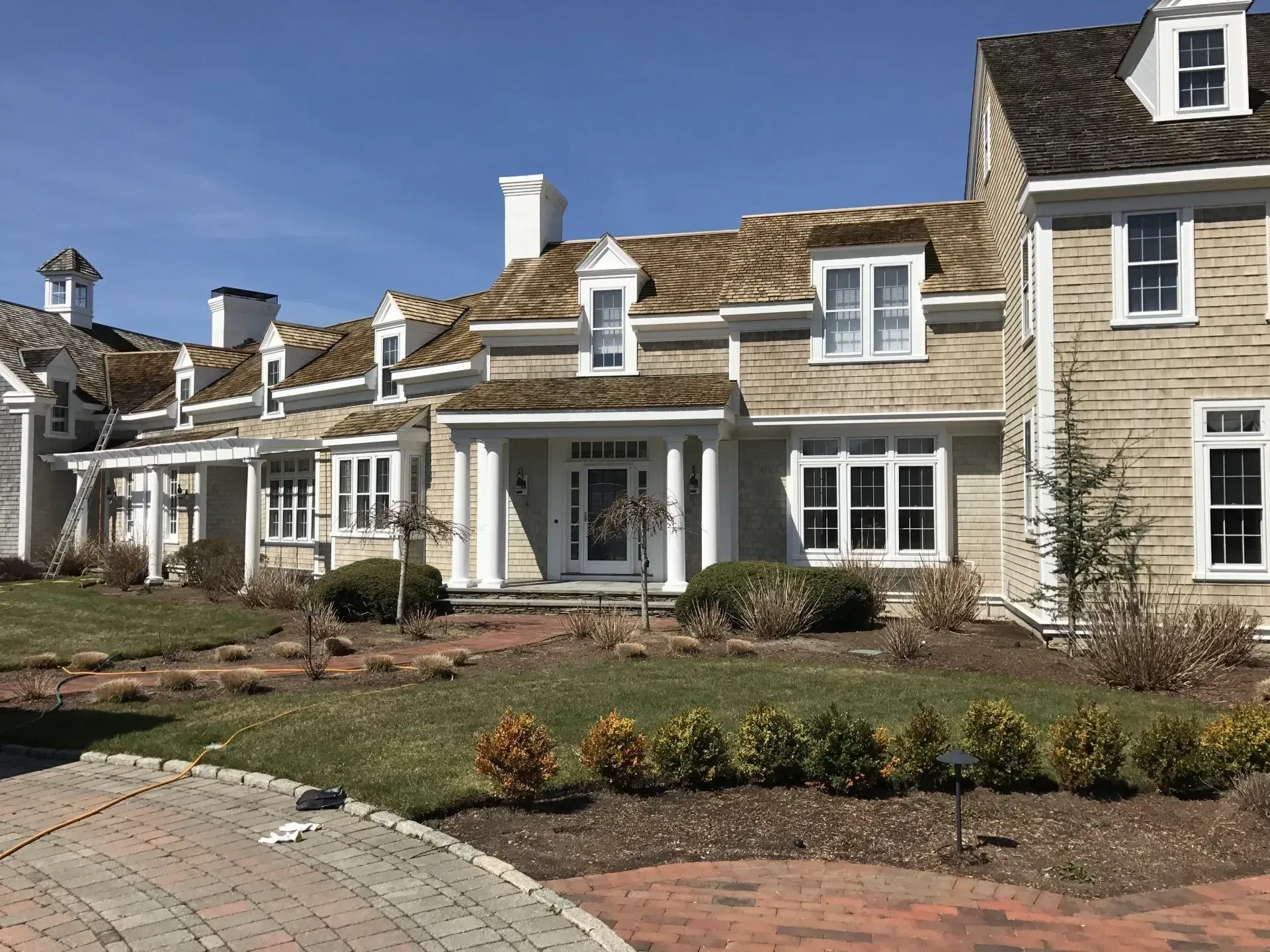 Large beige house with white trim, dormers, and a brick driveway.