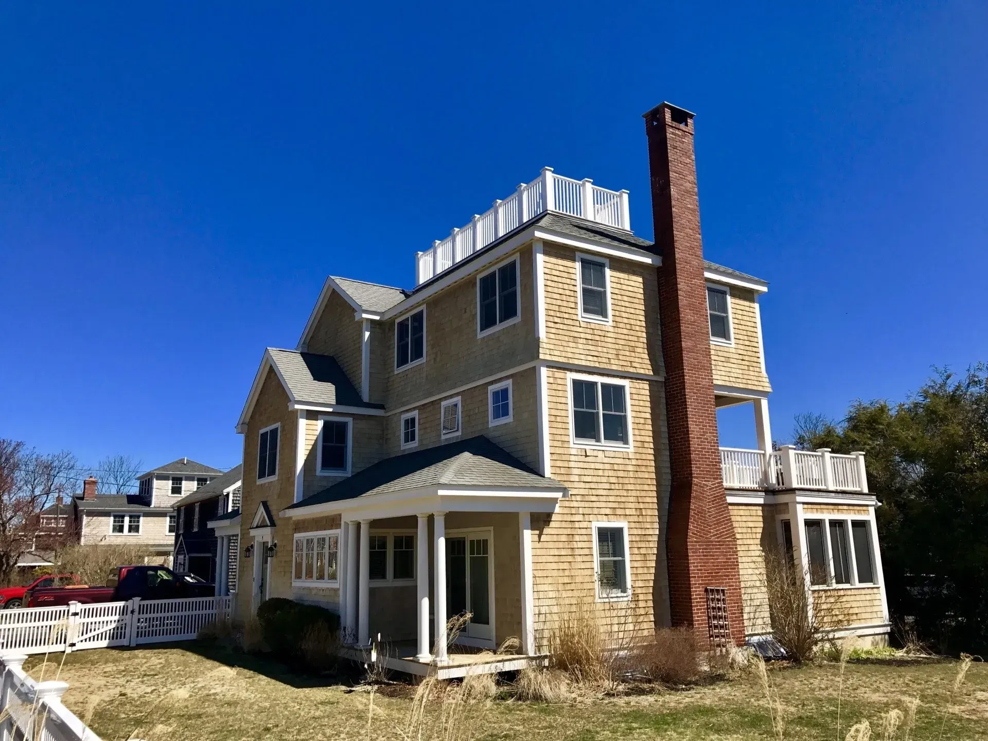 Three-story beige house with a brick chimney and white railing, under a blue sky.