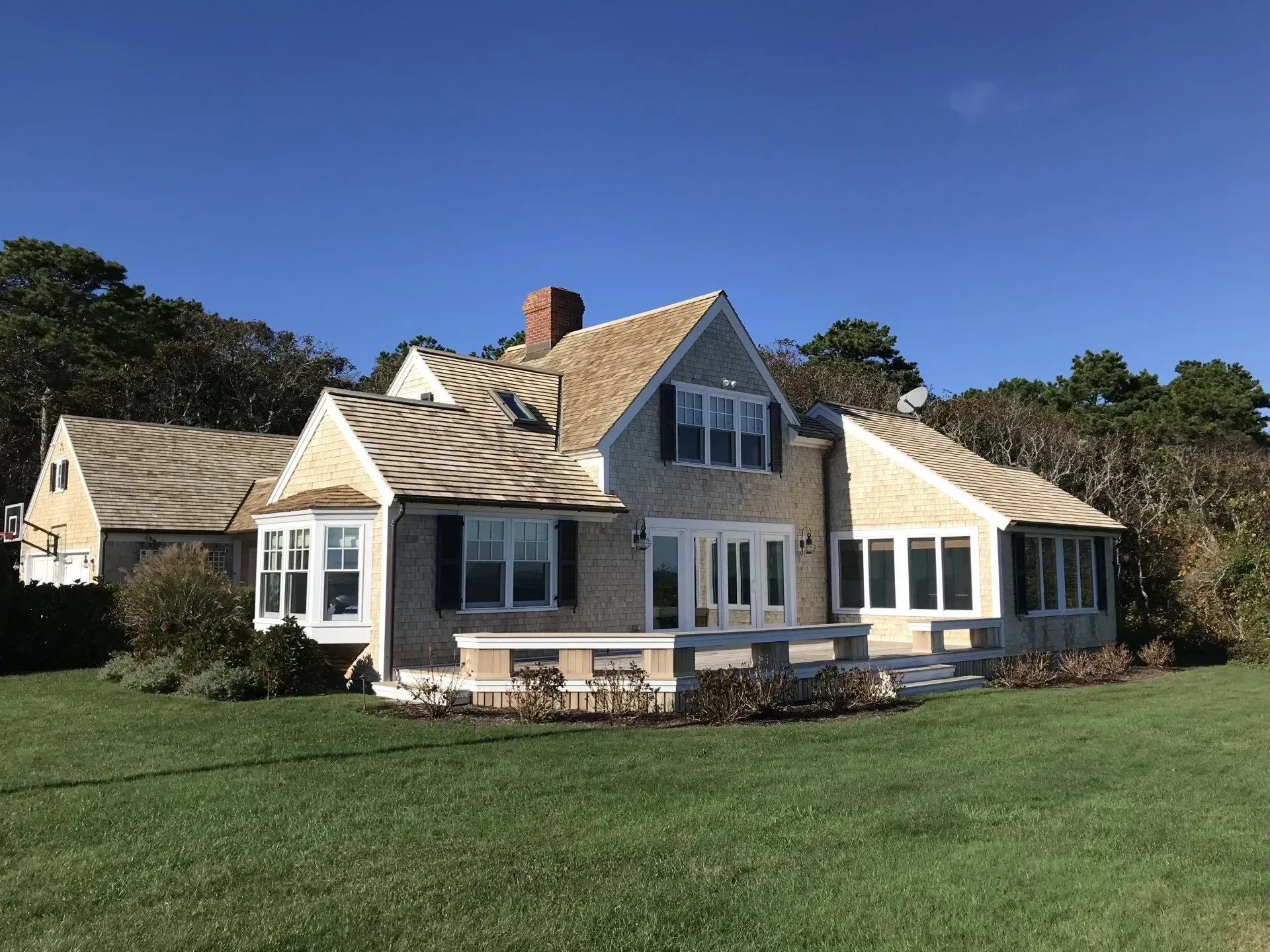 House with tan siding, beige roof, and dark shutters on a green lawn under a blue sky.