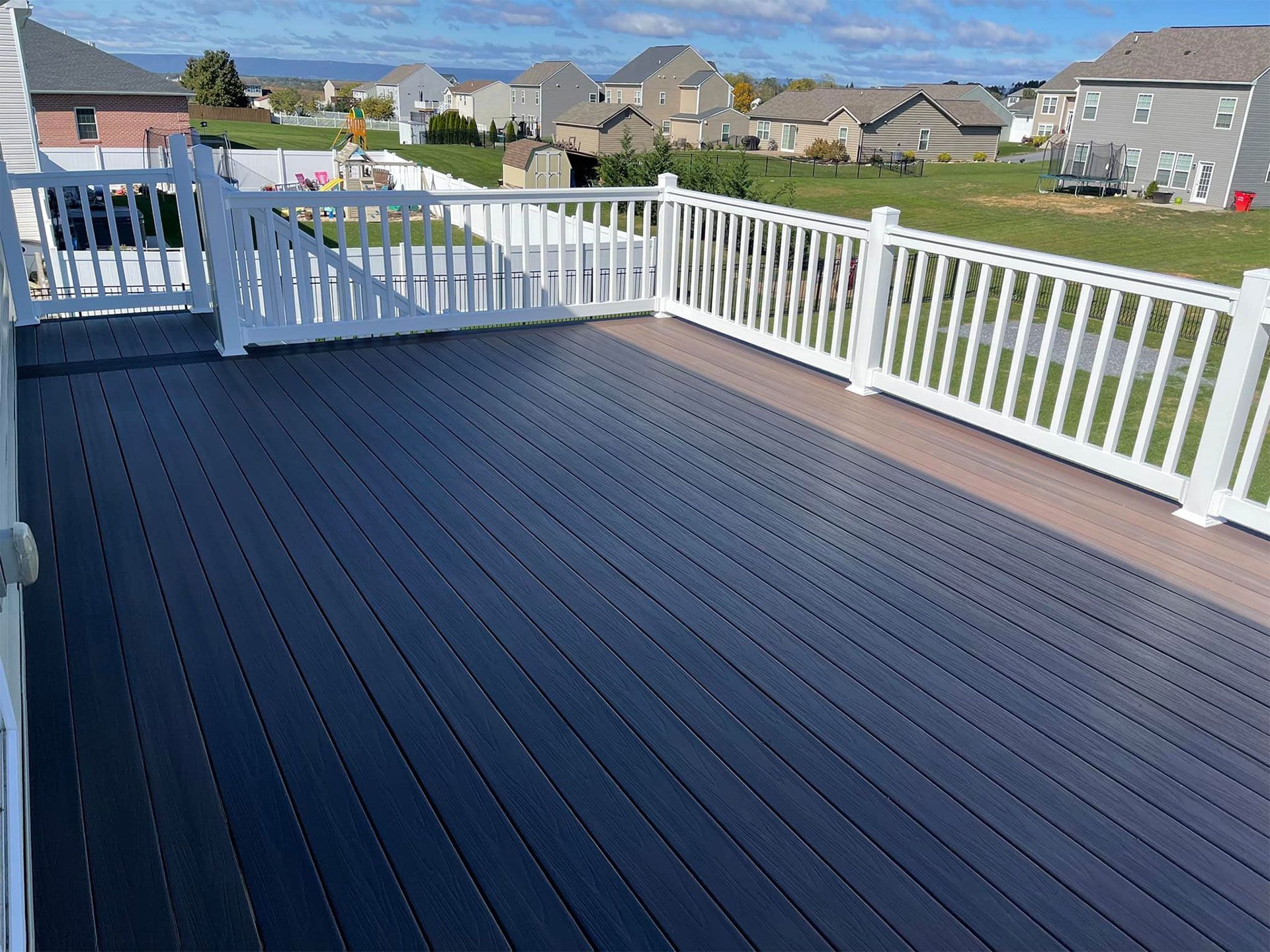 A large, dark composite deck with white railing overlooks a residential neighborhood on a sunny day.
