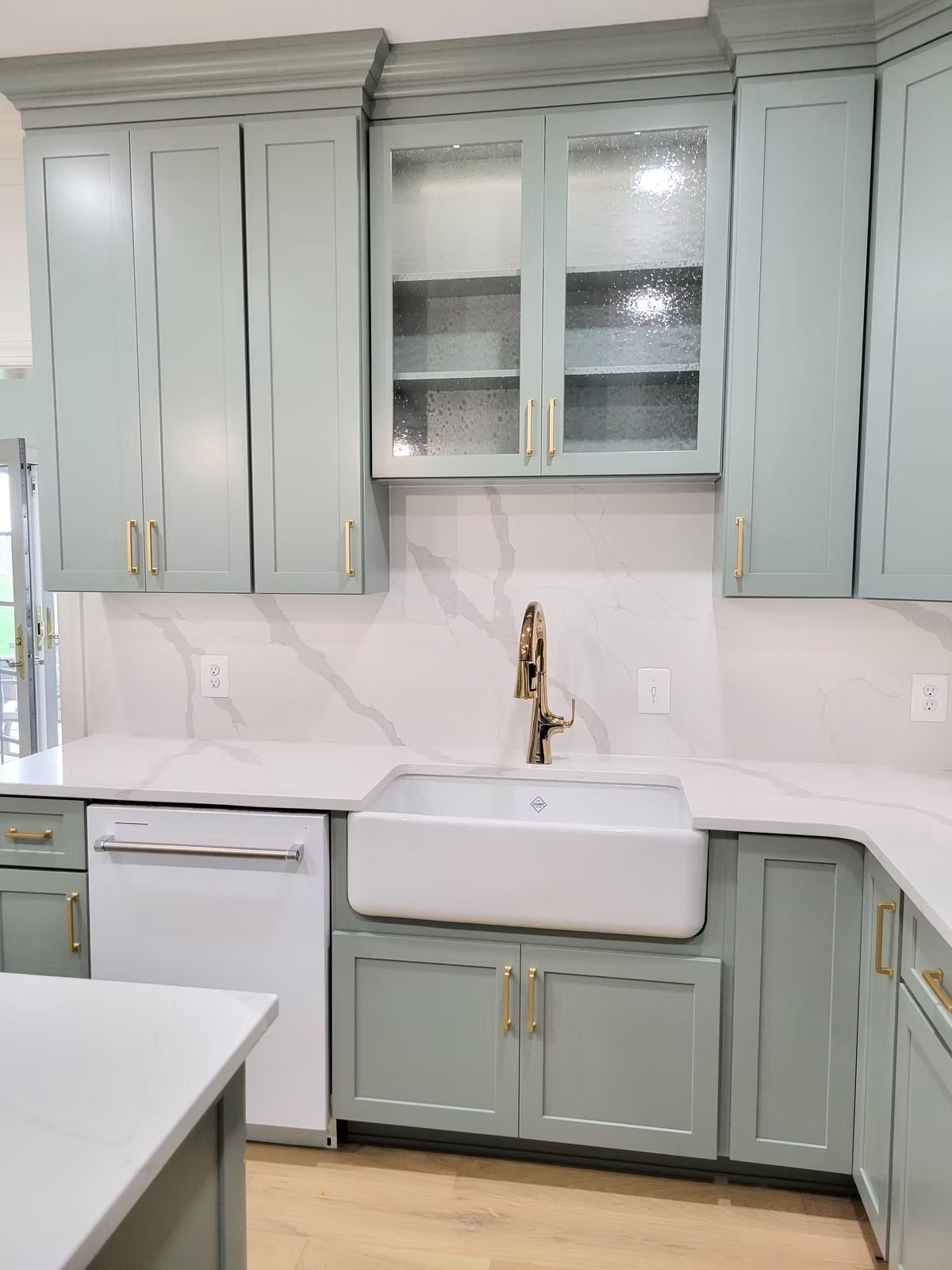 Light green kitchen cabinets with white countertops, sink, and dishwasher. Gold hardware.