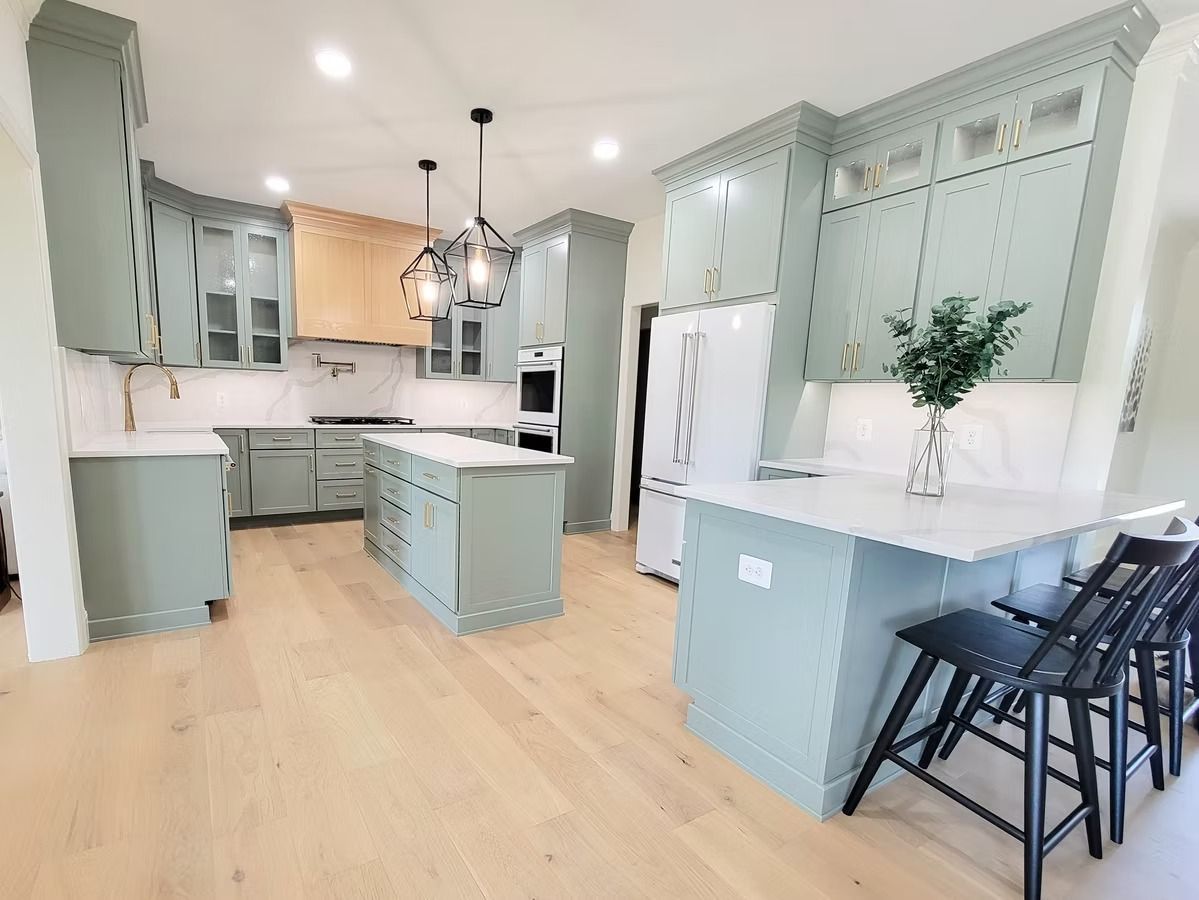 Spacious, light green kitchen with white countertops and wood flooring. Features include a large island and black bar stools.