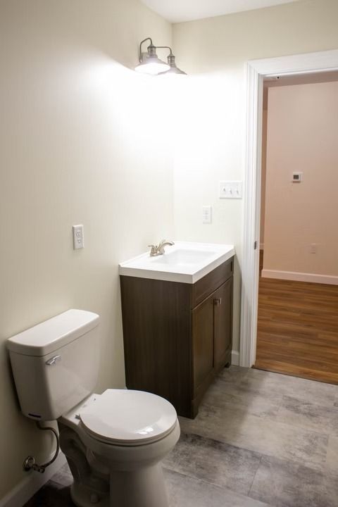 Bathroom with toilet, vanity, and open doorway, neutral color palette, concrete floor, overhead light.