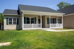 Beige ranch house with porch and dark roof, green shutters, and a green lawn.
