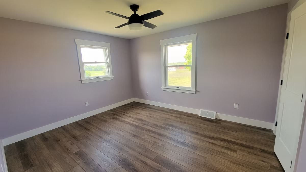 Empty bedroom with light purple walls, wood-look floor, and white trim. Ceiling fan and two windows.