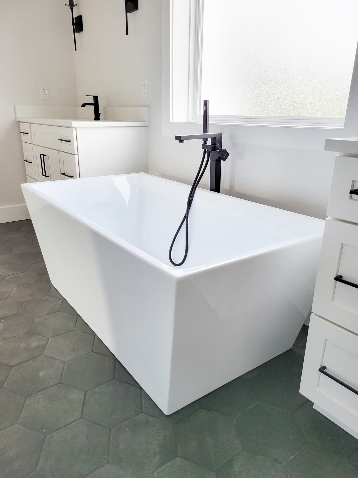 Modern white rectangular bathtub with black fixtures, set in a bathroom with dark gray hexagon tile flooring.