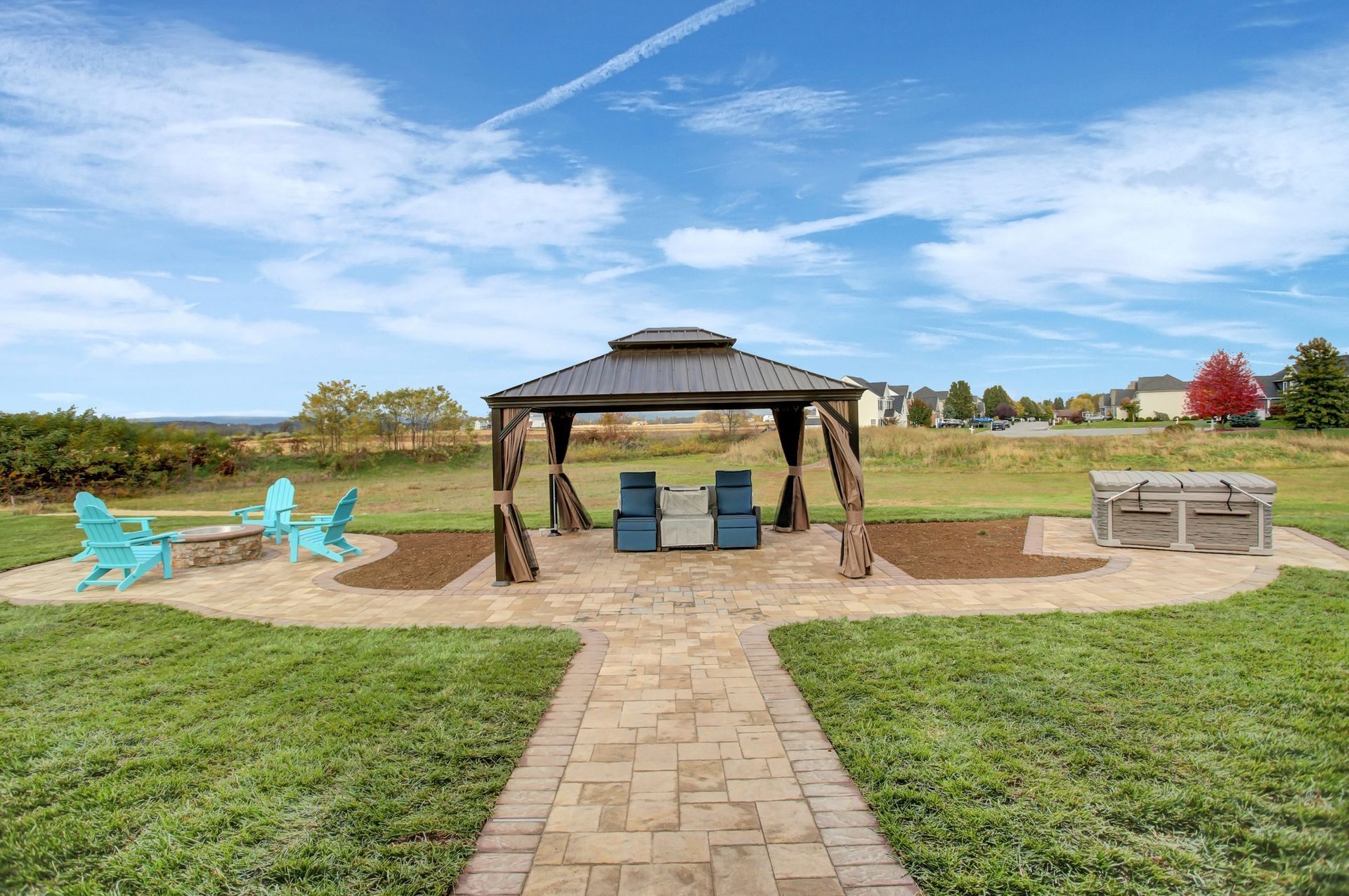 Outdoor patio with gazebo, seating, fire pit, and brick pathway on a grassy lawn under a blue sky.