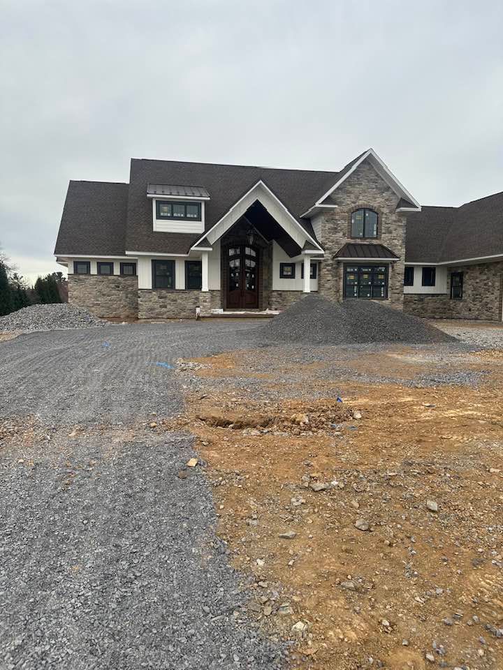 A large stone house under construction with a gravel driveway and cloudy sky.