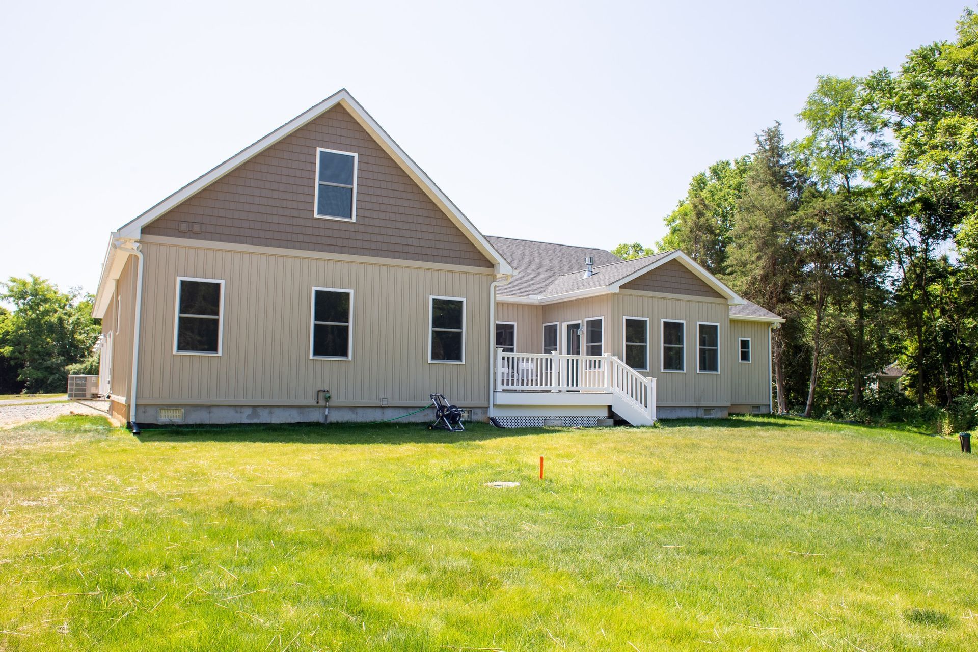 Tan house with brown trim and windows, white deck, green grass, and trees under a clear blue sky.