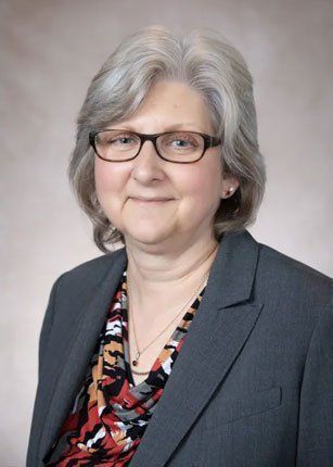 Woman with gray hair and glasses wearing a blazer and patterned blouse, smiling.
