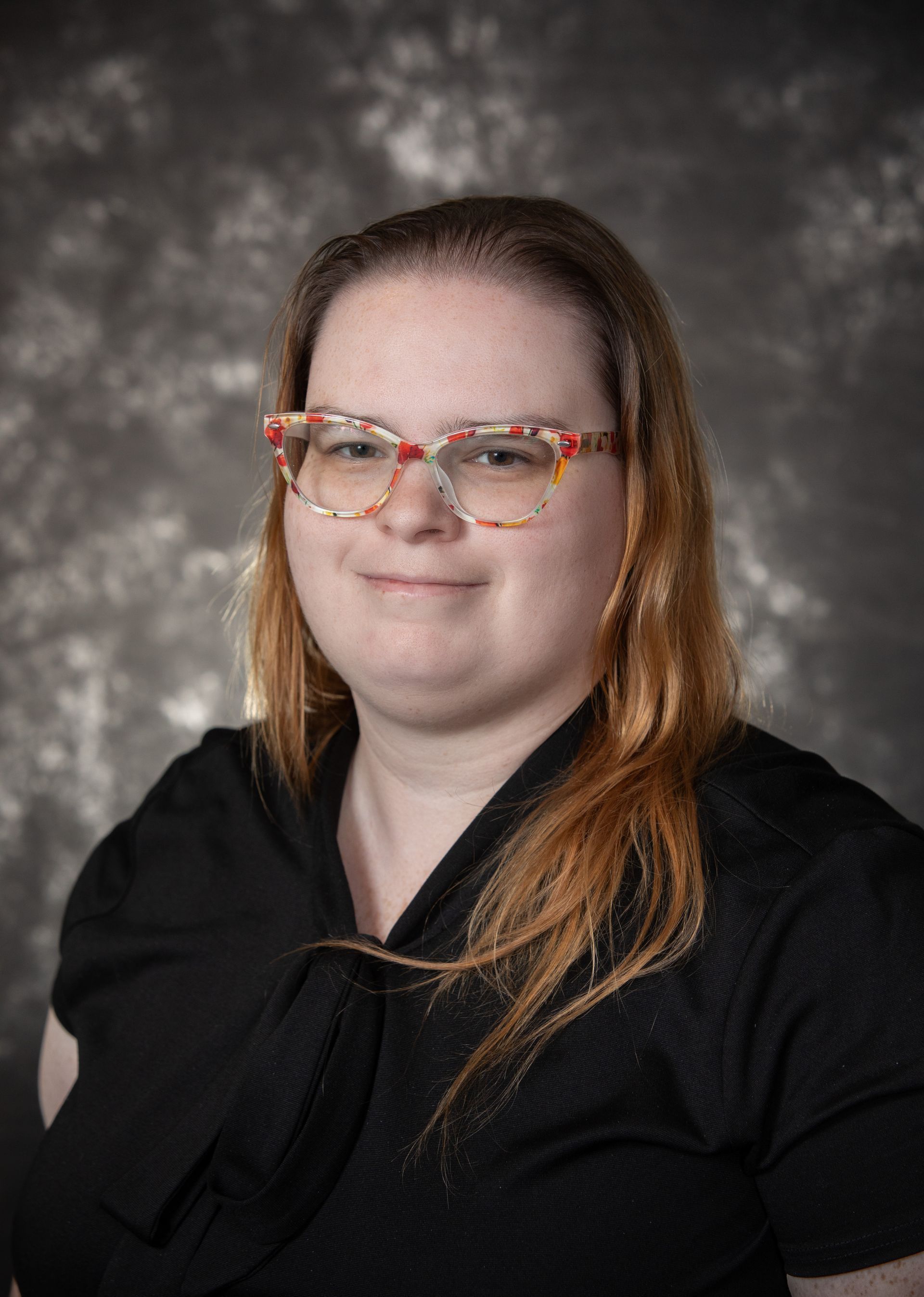 Woman with glasses, wearing a black top, smiling. Grey backdrop.