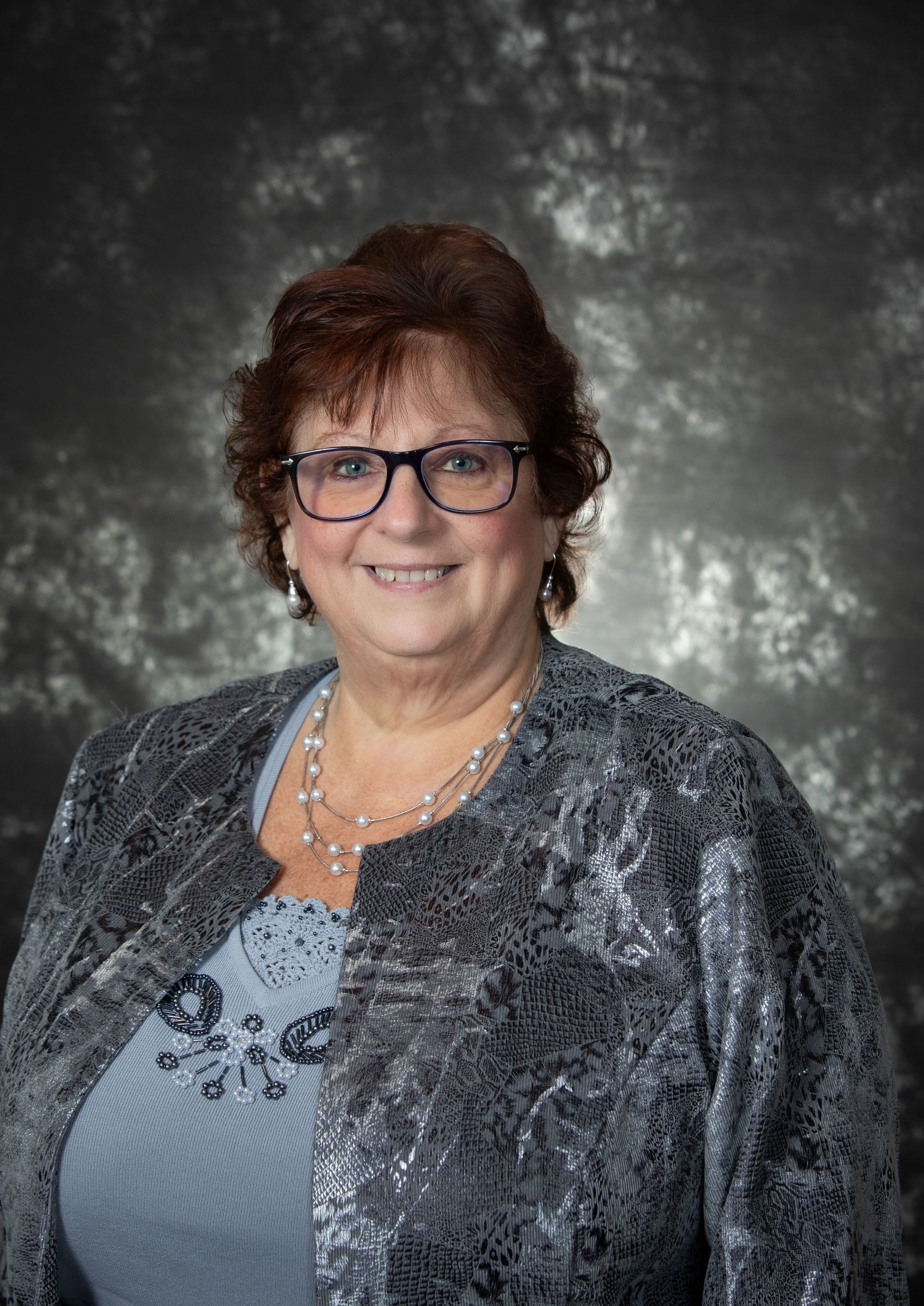 Woman with glasses, brown hair, smiling, wearing a gray sequined jacket and blue top, against a mottled background.