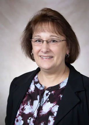 Woman with glasses smiling, wearing patterned shirt and black blazer, headshot.