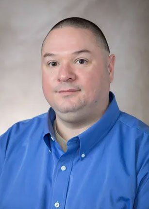 Man in a blue shirt smiles at the camera; light skin, short dark hair, indoors.