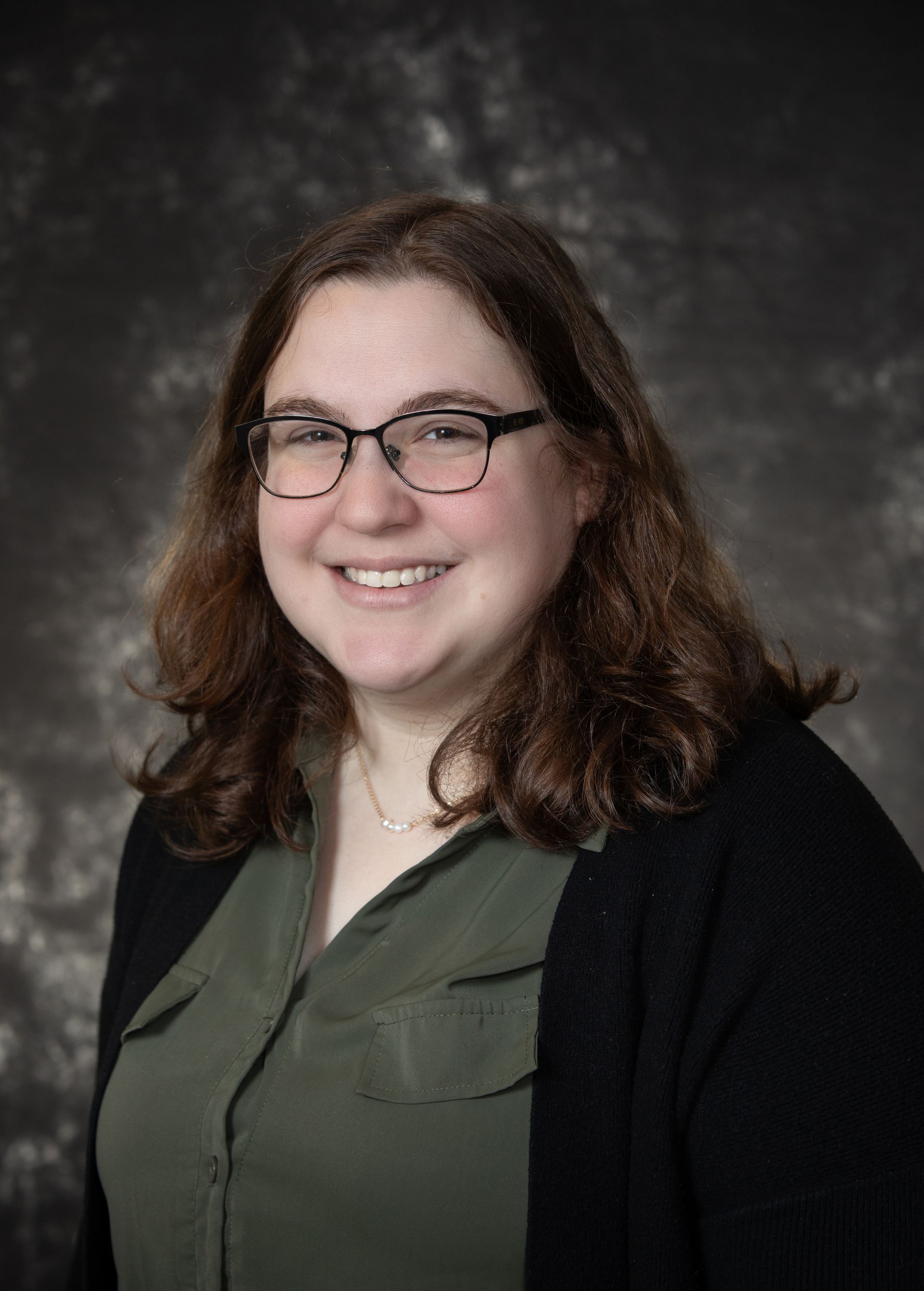 Woman with glasses smiles, wearing green shirt, black cardigan; dark background.