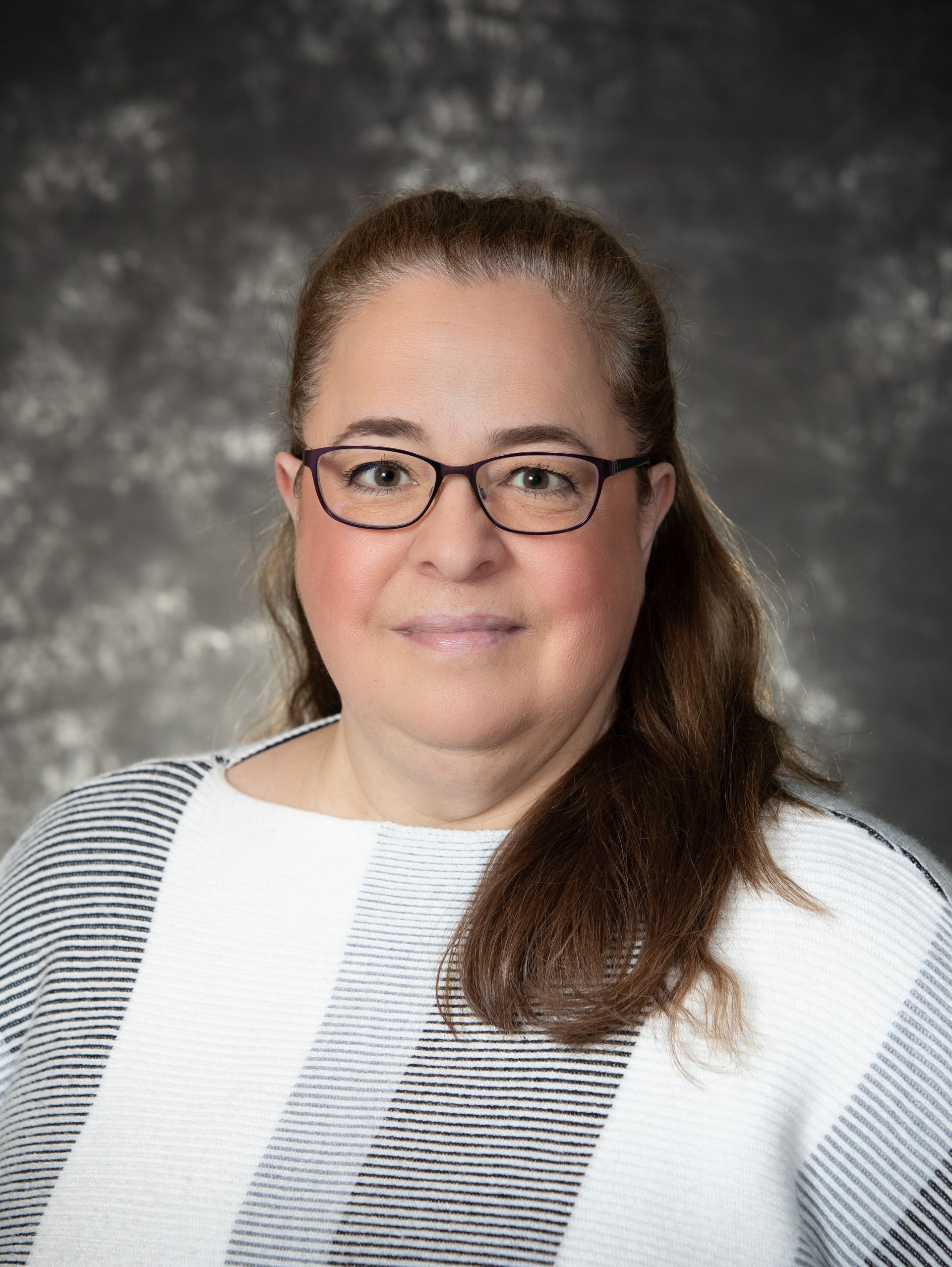 Woman with glasses, looking forward. Wearing a black and white striped sweater. Gray background.