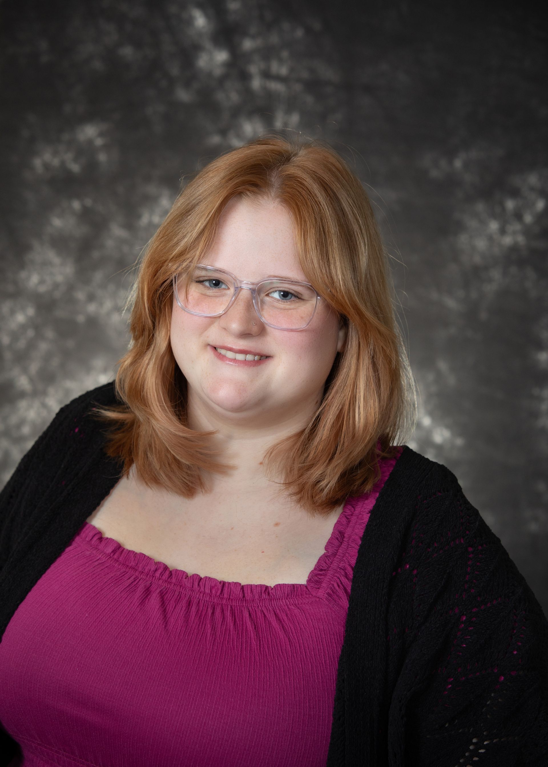 Young woman with red hair and glasses, wearing a pink shirt and black cardigan, smiles in front of a gray backdrop.