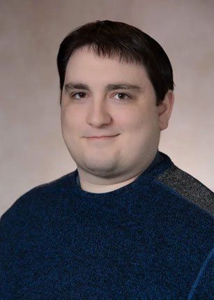 Man with dark hair, wearing a blue sweater, smiling at the camera. Neutral background.