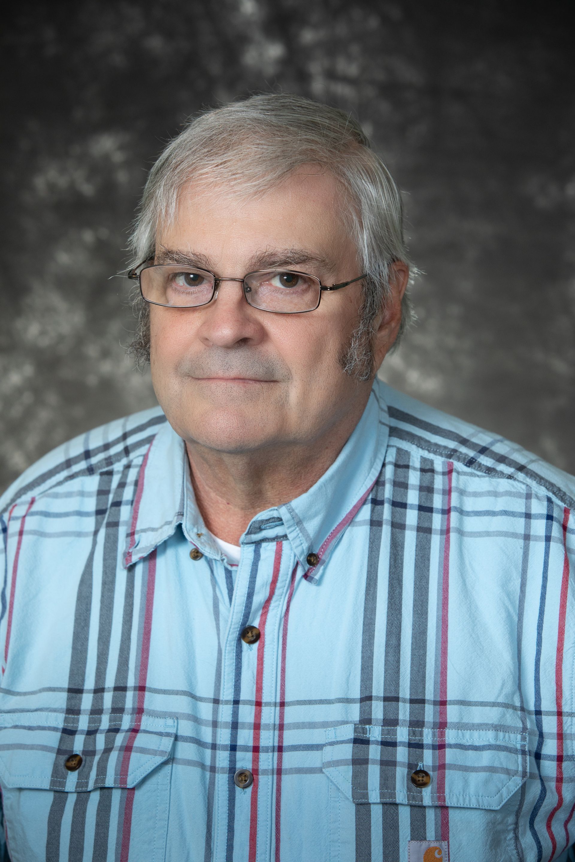 Man with glasses wearing blue plaid shirt, looking at the viewer, with a blurred background.