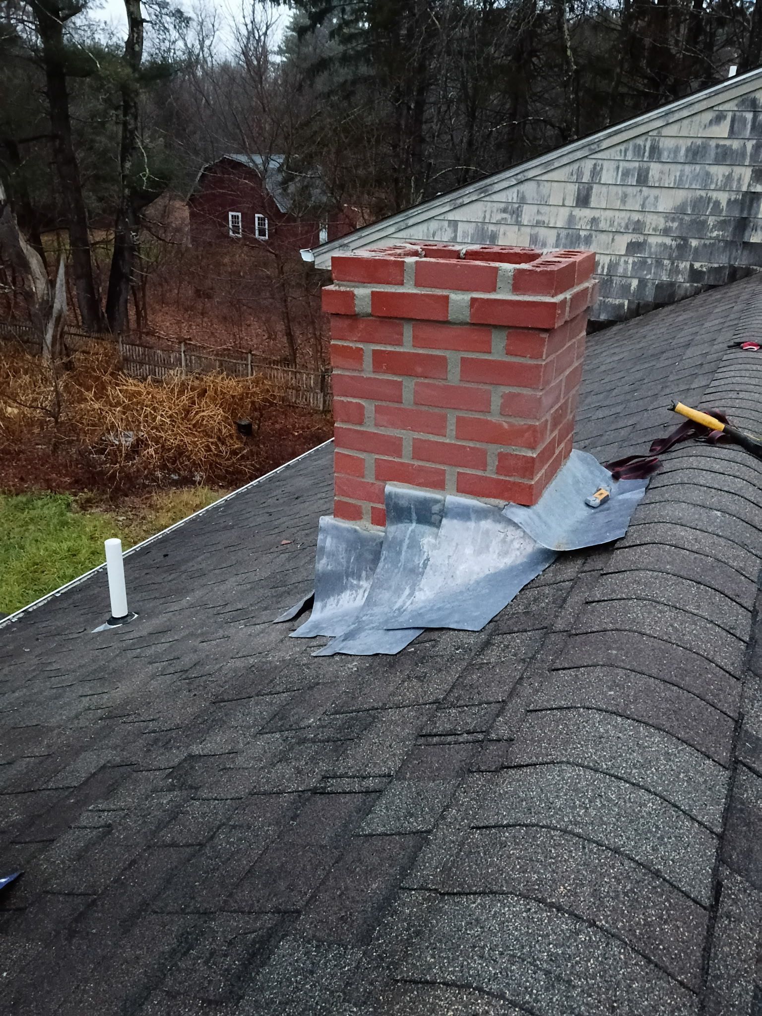 Red brick chimney on a dark shingle roof with flashing; autumn foliage in the background.