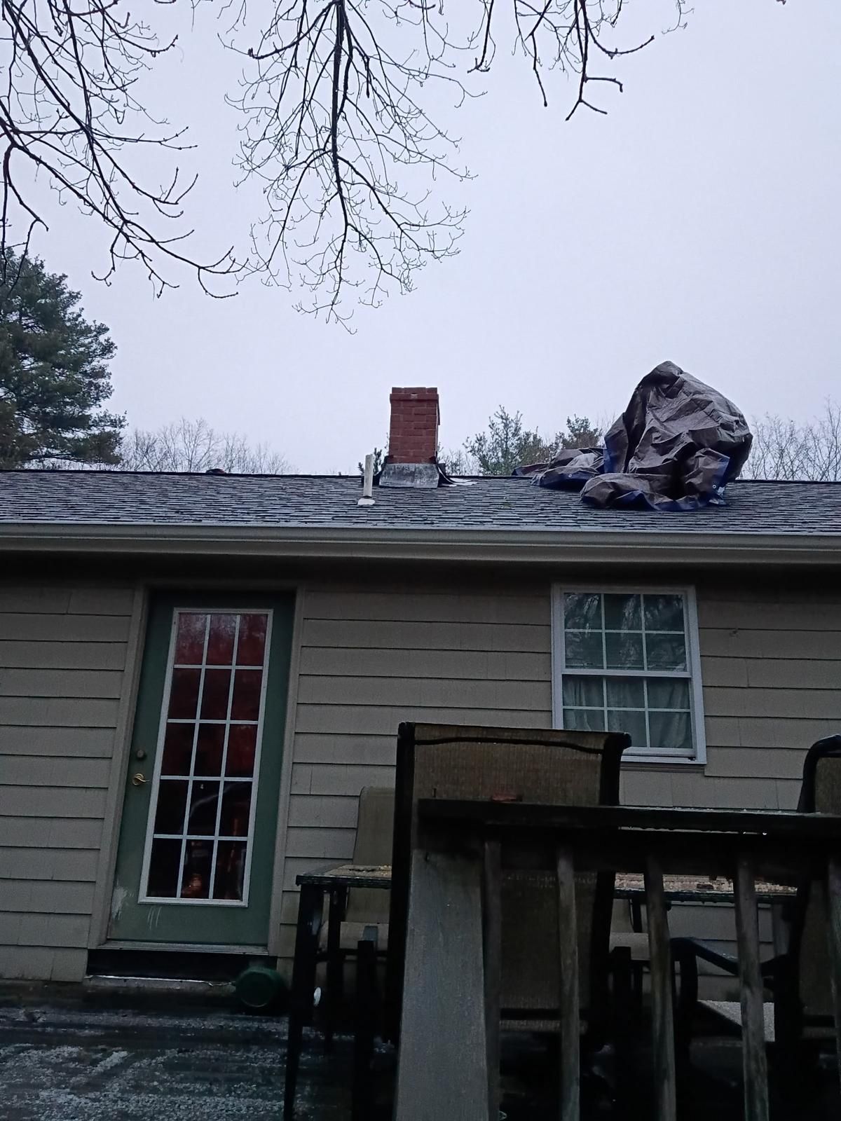 A brick chimney and tarp on a snow-covered roof, cloudy sky above a beige house.