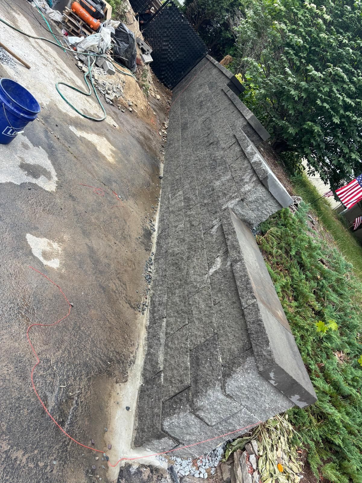 Concrete retaining wall next to paved area, blue bucket, and green hose visible.