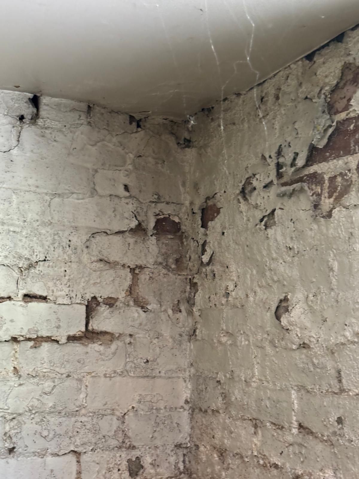 Corner of a brick wall, painted white, with visible cracks and peeling paint. A ceiling meets the wall.