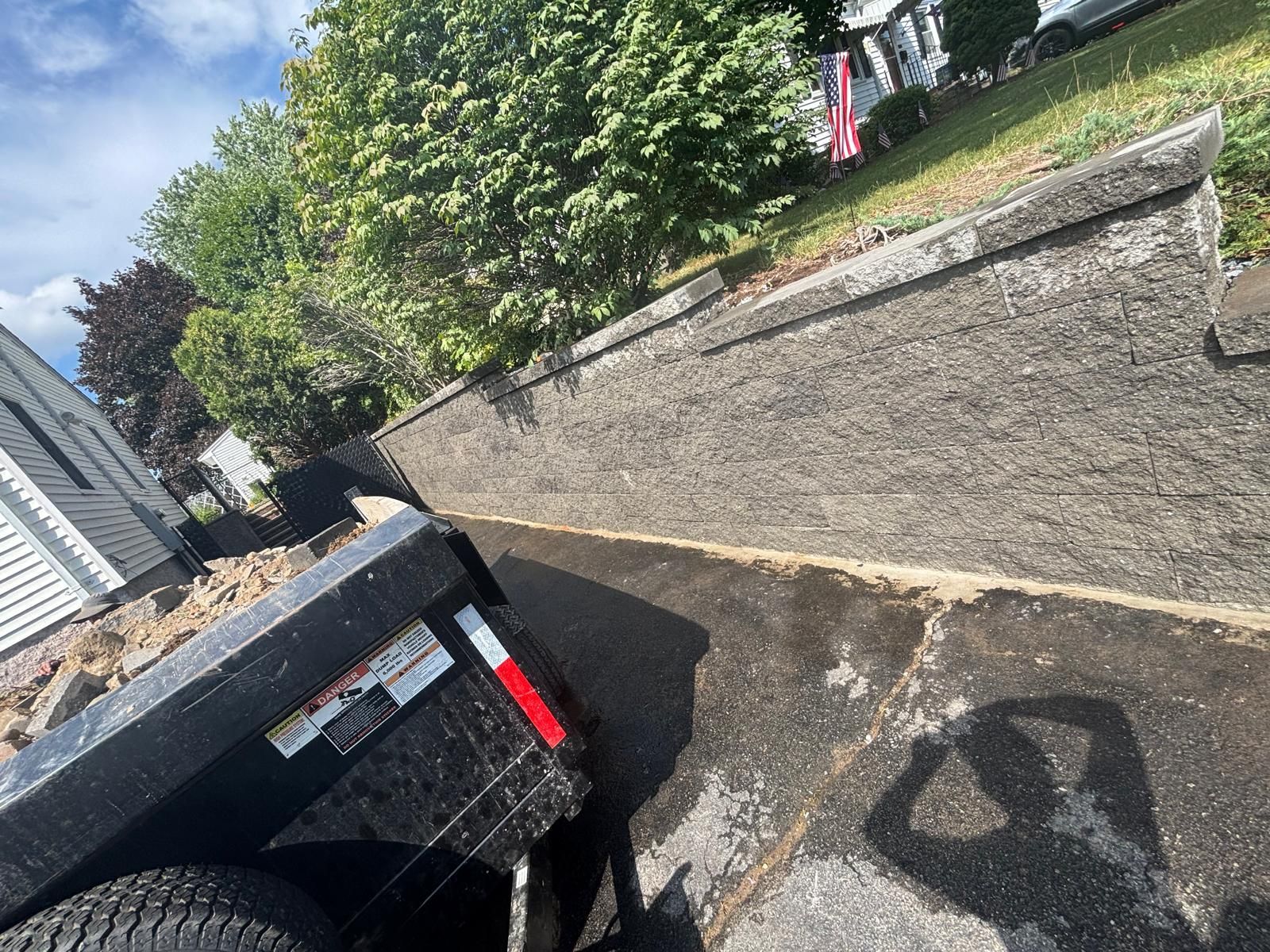 Black trailer parked next to a concrete retaining wall, a house and foliage in the background.