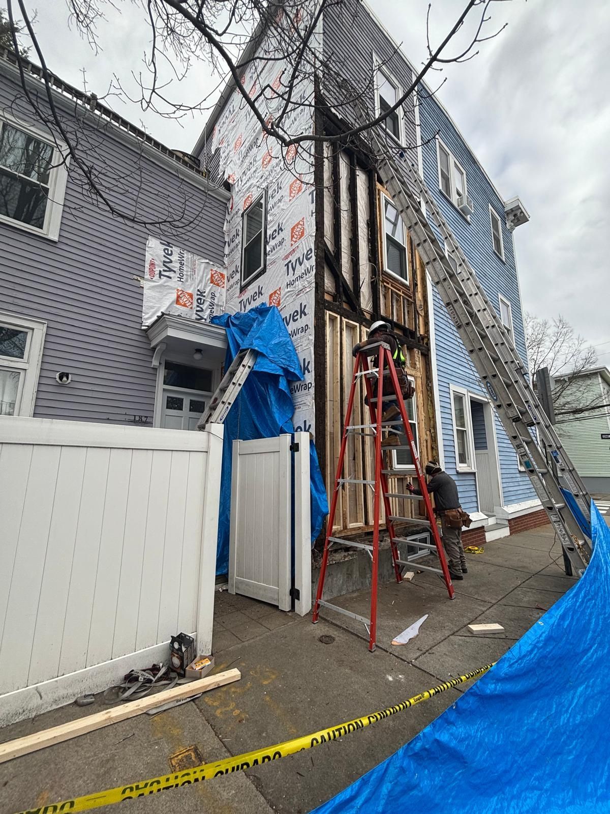 House exterior undergoing construction. A worker on a ladder, Tyvek wrap, blue tarp, and a tall ladder visible.