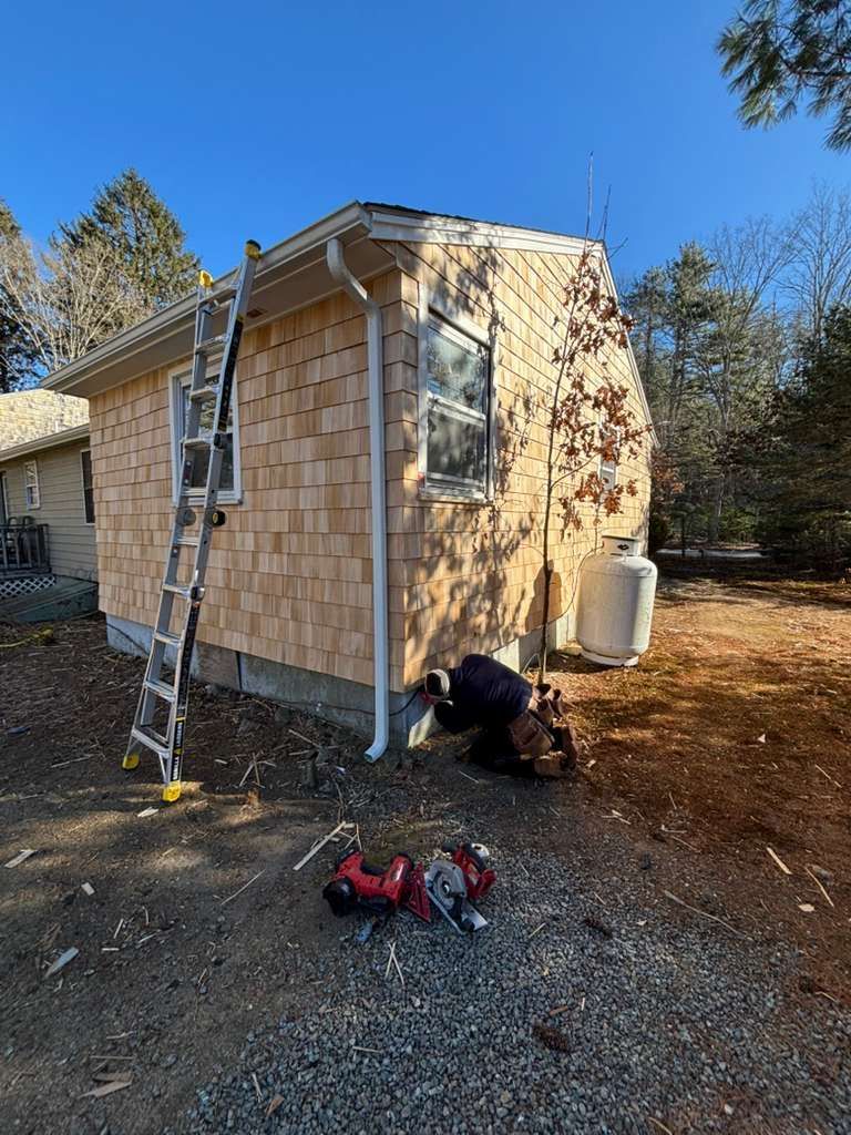 Construction of a small building; wooden shingles being installed. A person works near the ground.
