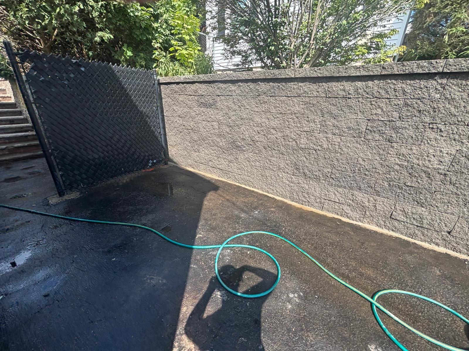 A black fence and concrete wall being hosed down with a green hose on a sunny day.