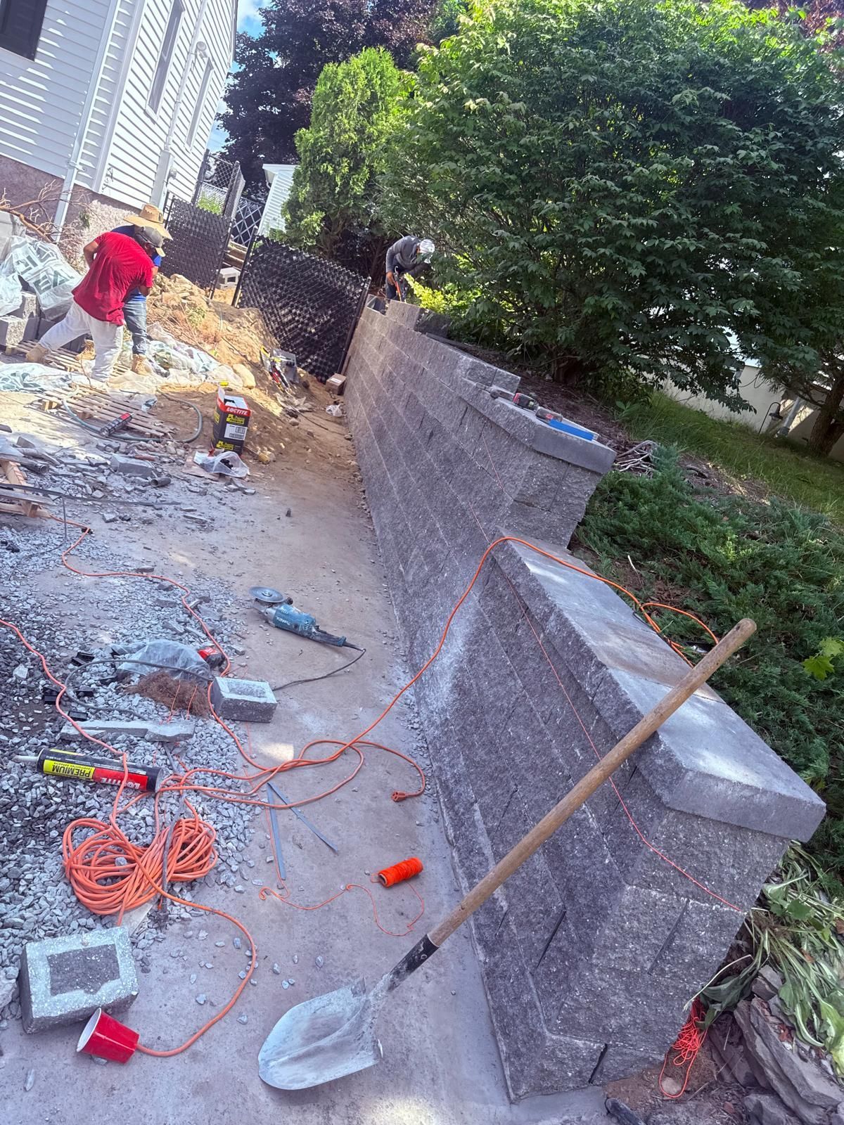 Construction site with a retaining wall being built. Tools and materials are scattered, workers are present.