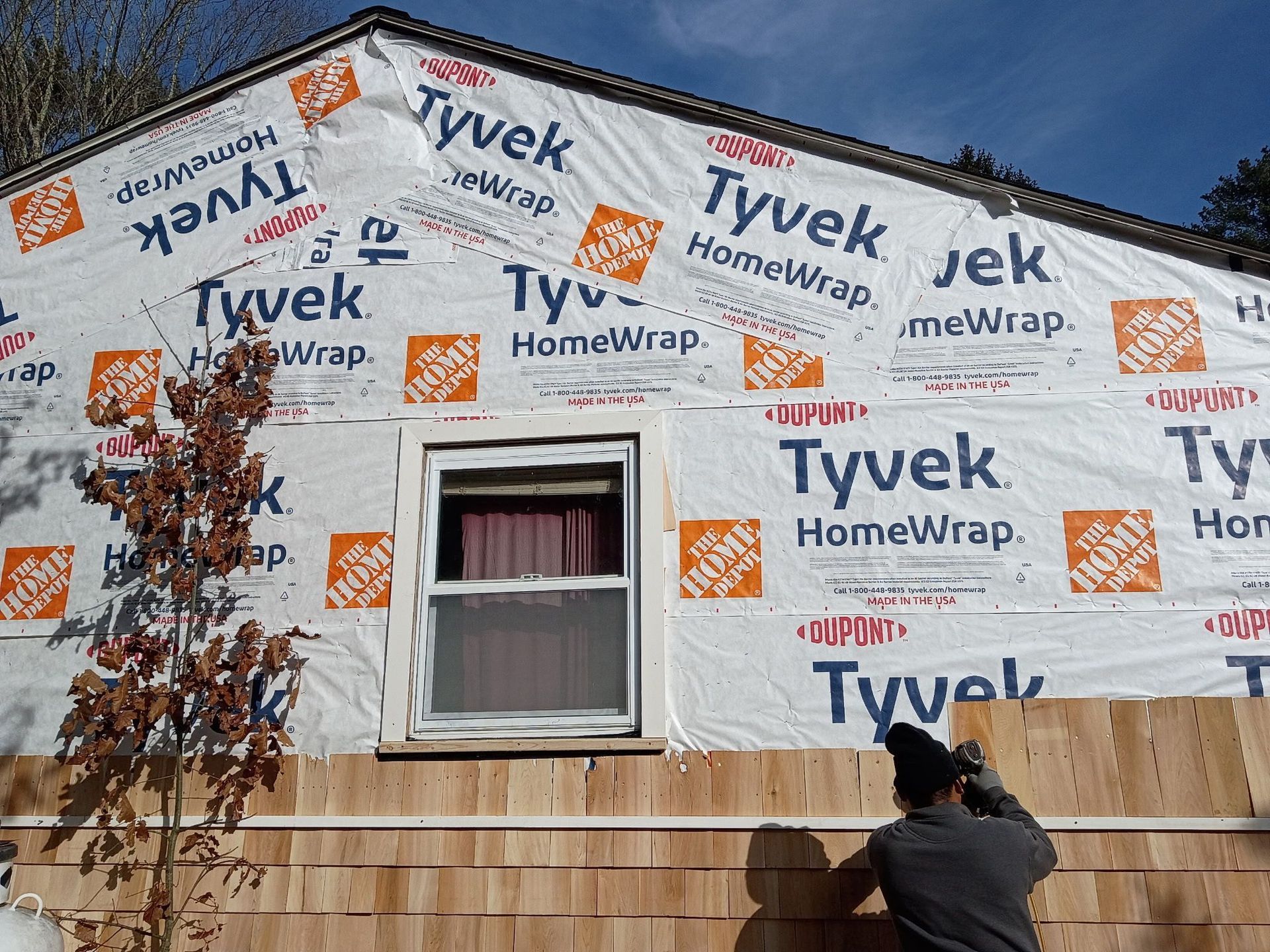 House under construction, covered with Tyvek and cedar shingles; a person is working on the siding.