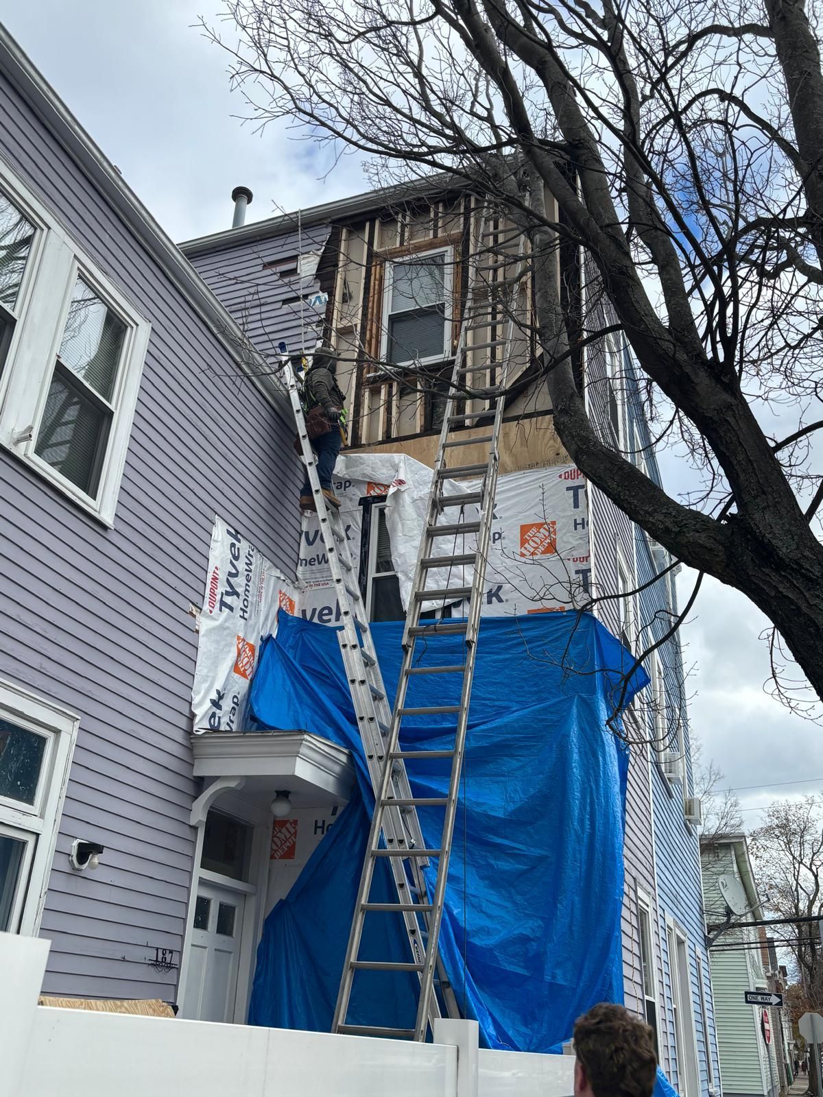 Building exterior under construction; ladders, blue tarp, exposed wood, worker.