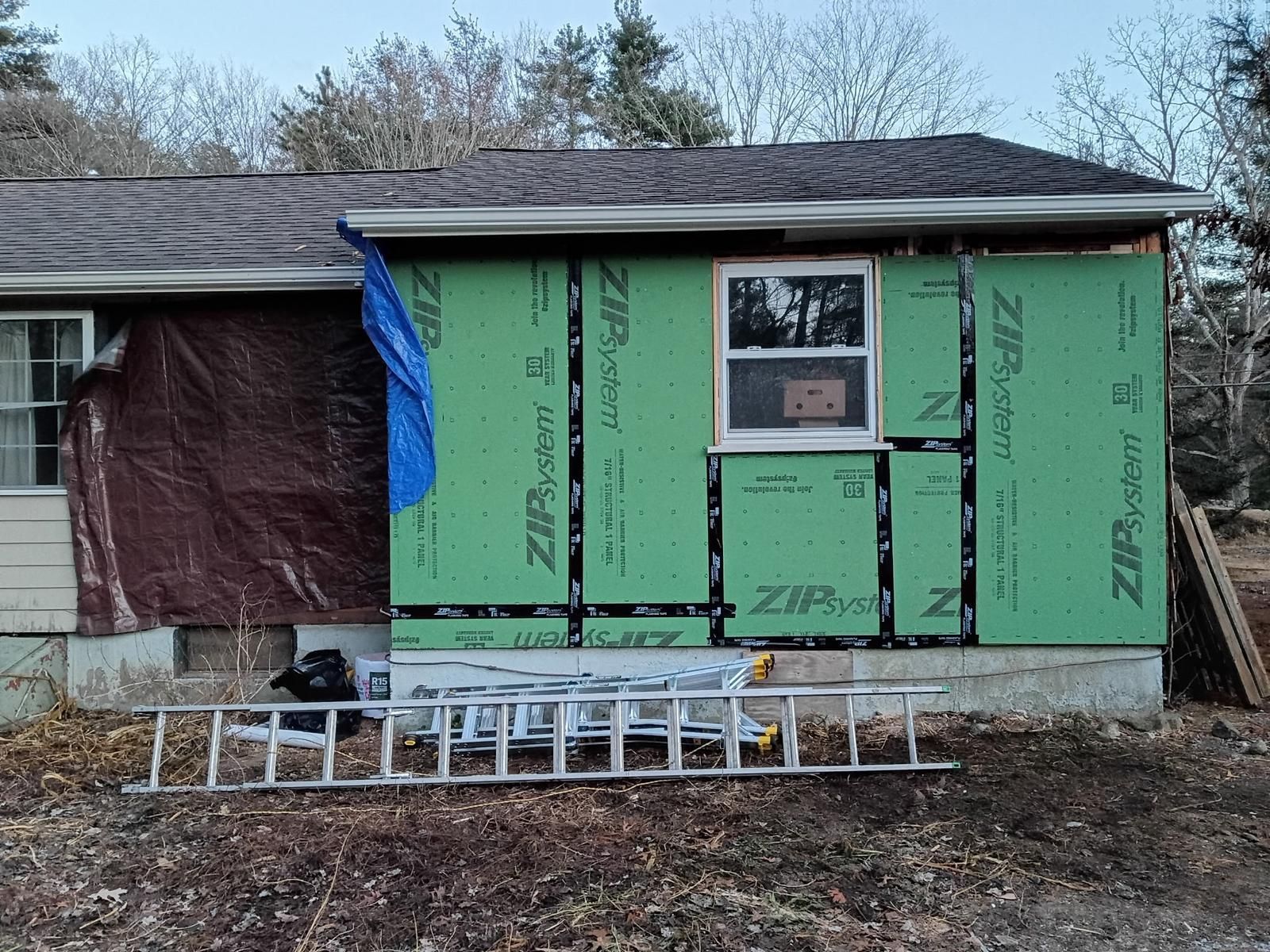 House under construction; green sheathing covers a new addition's frame, window installed. A tarp covers existing siding.