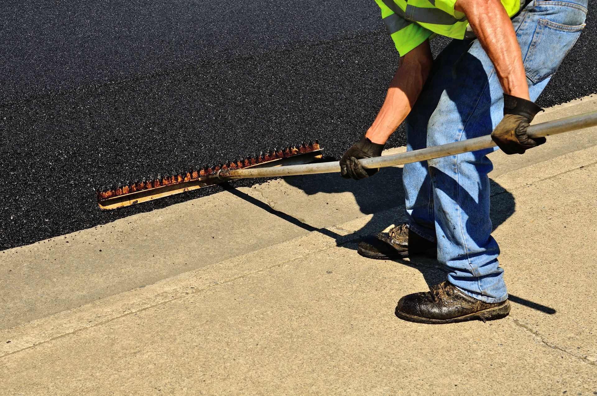 Construction worker using a rake to spread hot asphalt evenly along the edge of a sidewalk.