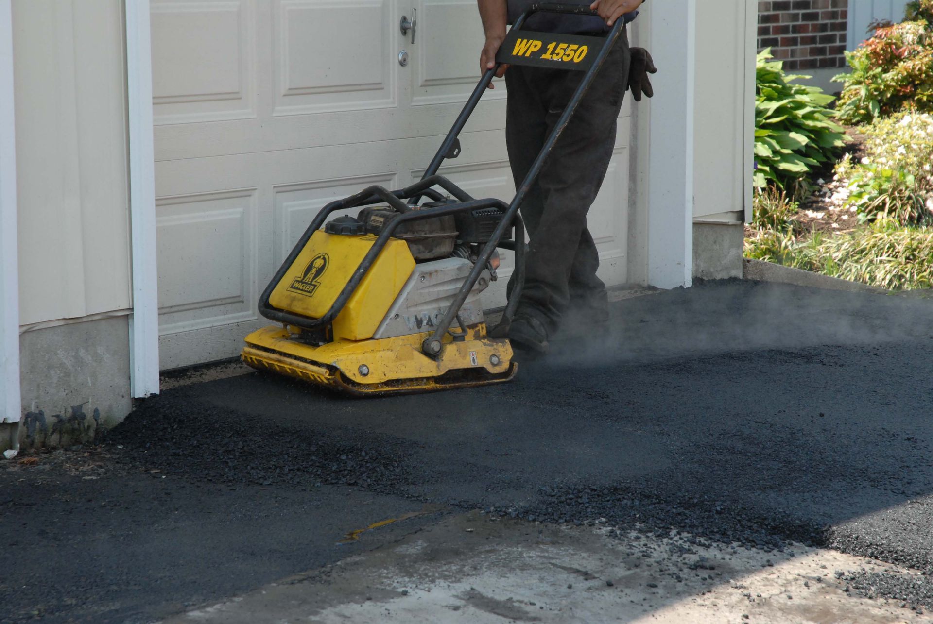Worker using a plate compactor to press hot asphalt on a residential driveway near a garage.