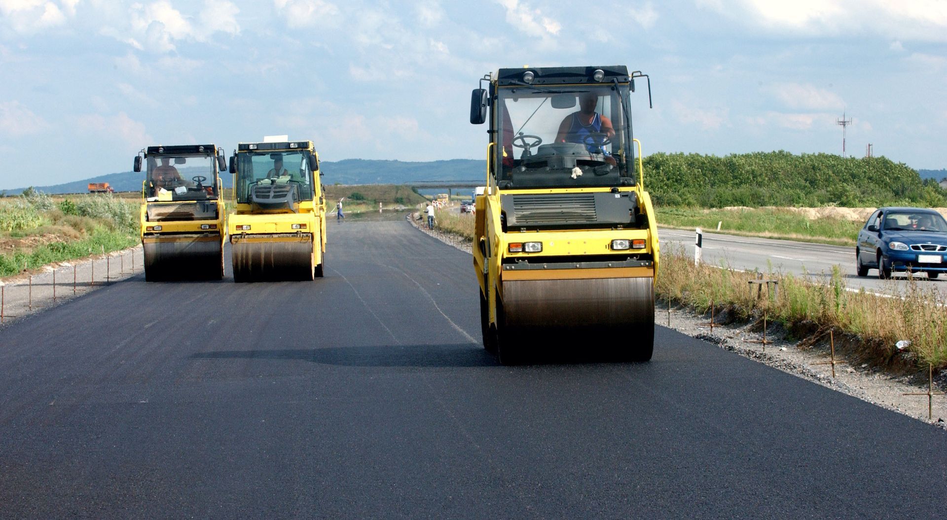Three steamrollers compacting fresh asphalt on a rural highway under construction.