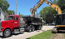 Red dump truck loading debris beside a yellow excavator on a residential street.