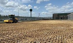 Yellow tractor grading a dirt lot near a water tower and industrial buildings under a cloudy sky.