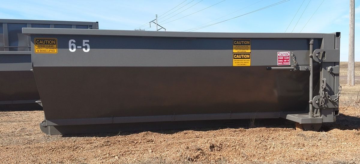 A large, gray metal dumpster outdoors on brown ground under a blue sky.  It has yellow warning stickers.