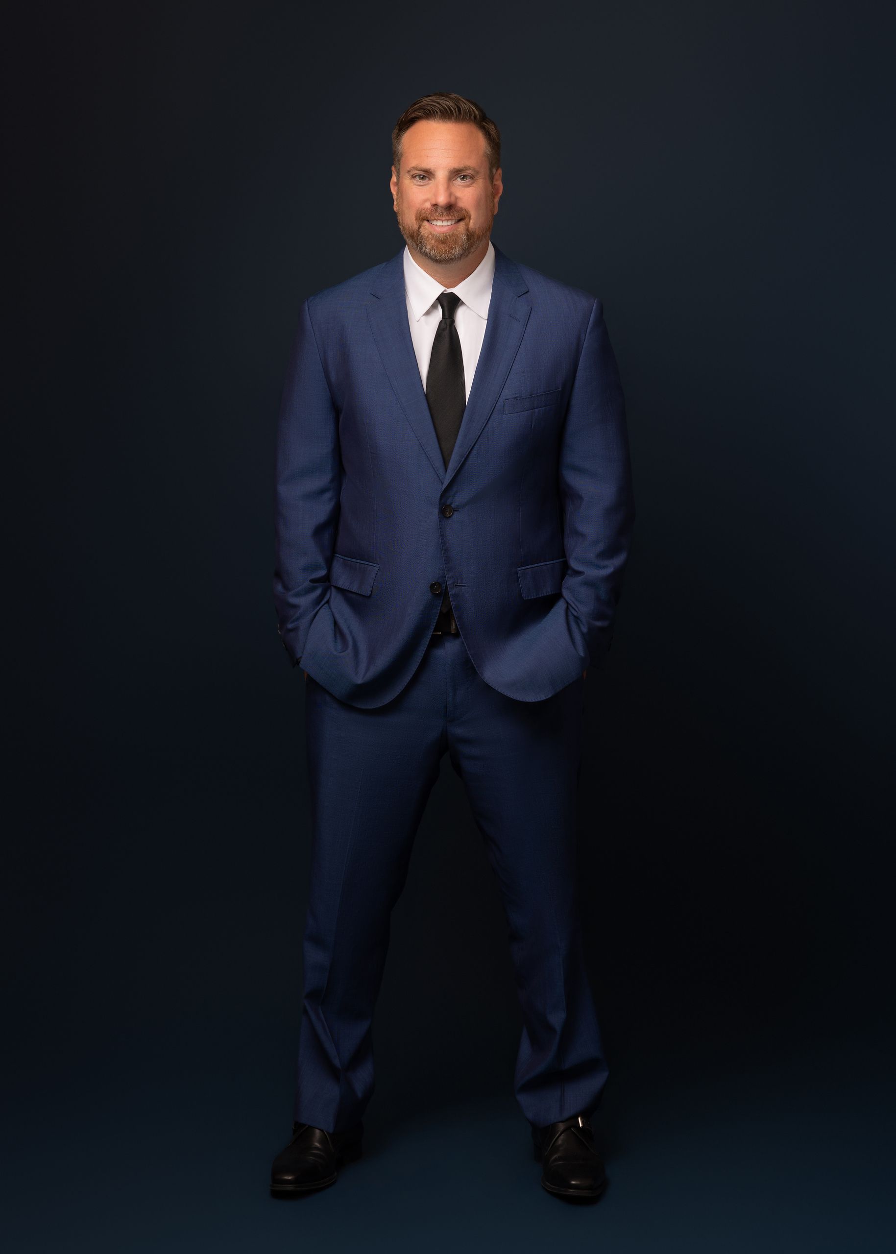 Man in blue suit, hands in pockets, smiling against a dark blue background.