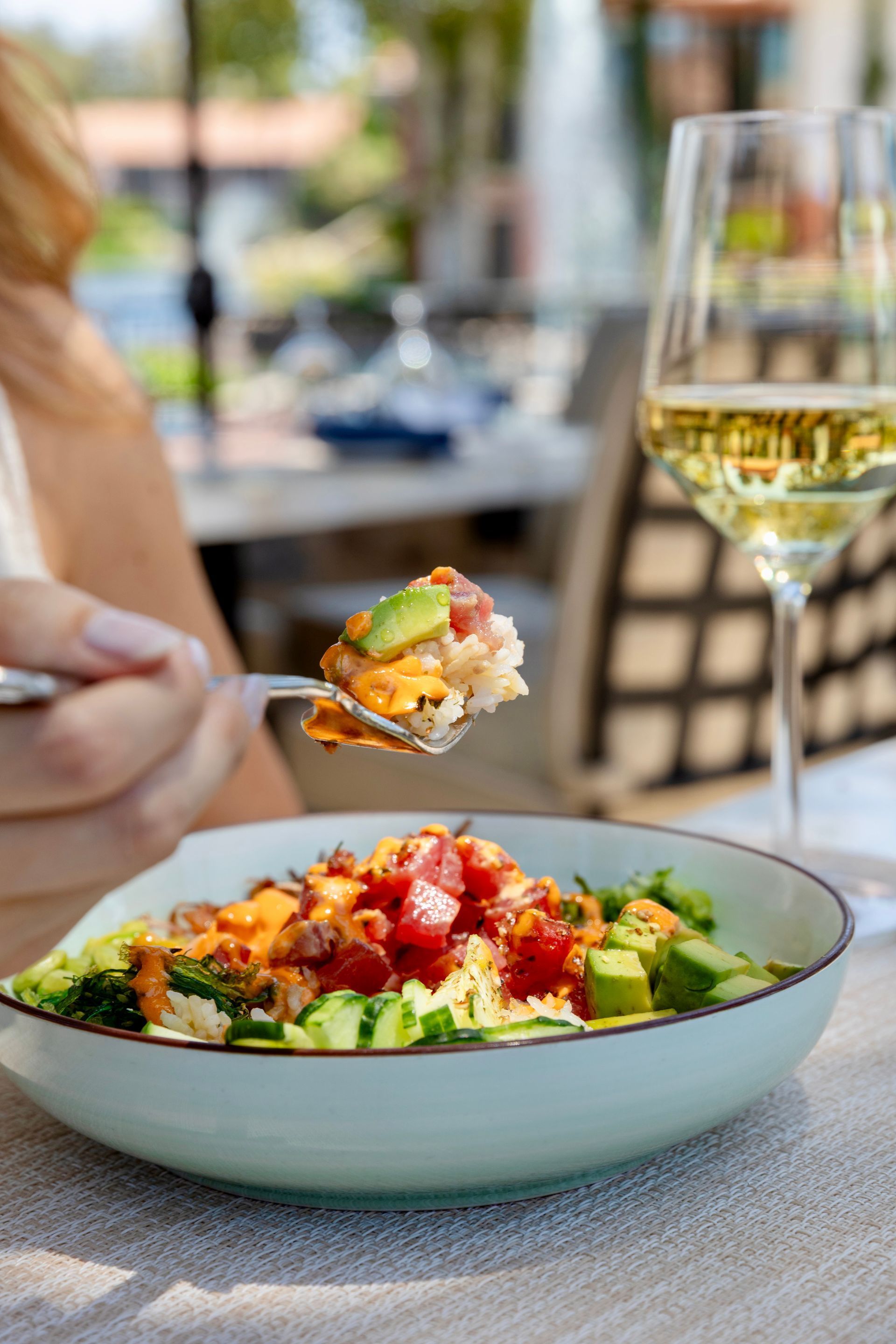 A plate of colorful salad beside a glass of white wine on a restaurant table, with a hand holding a forkful of food.