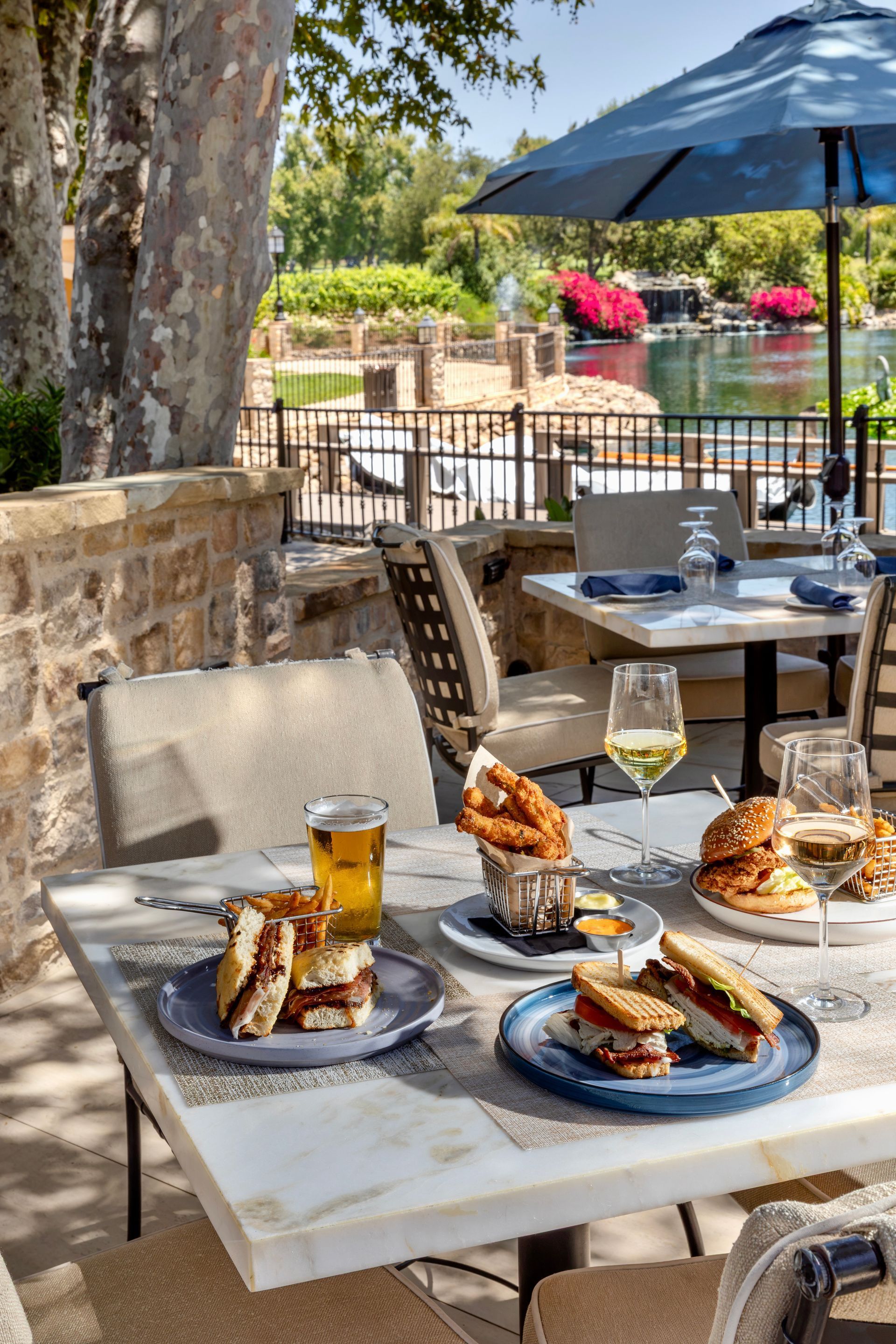 Outdoor café table with sandwiches, drinks, and blue umbrellas by a stone patio and water
