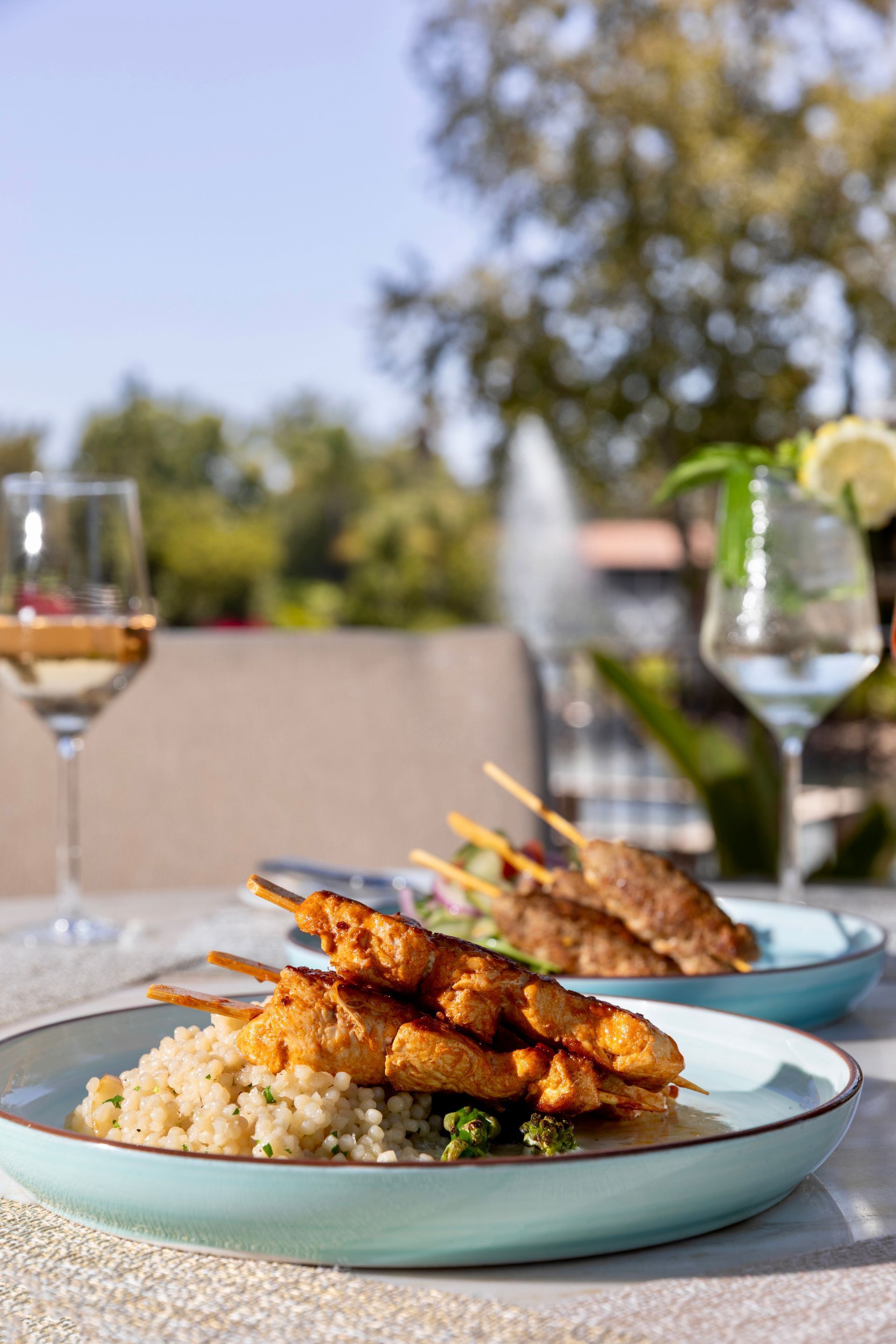 Outdoor table with plates of grilled skewers over rice, with wine glasses in the background.