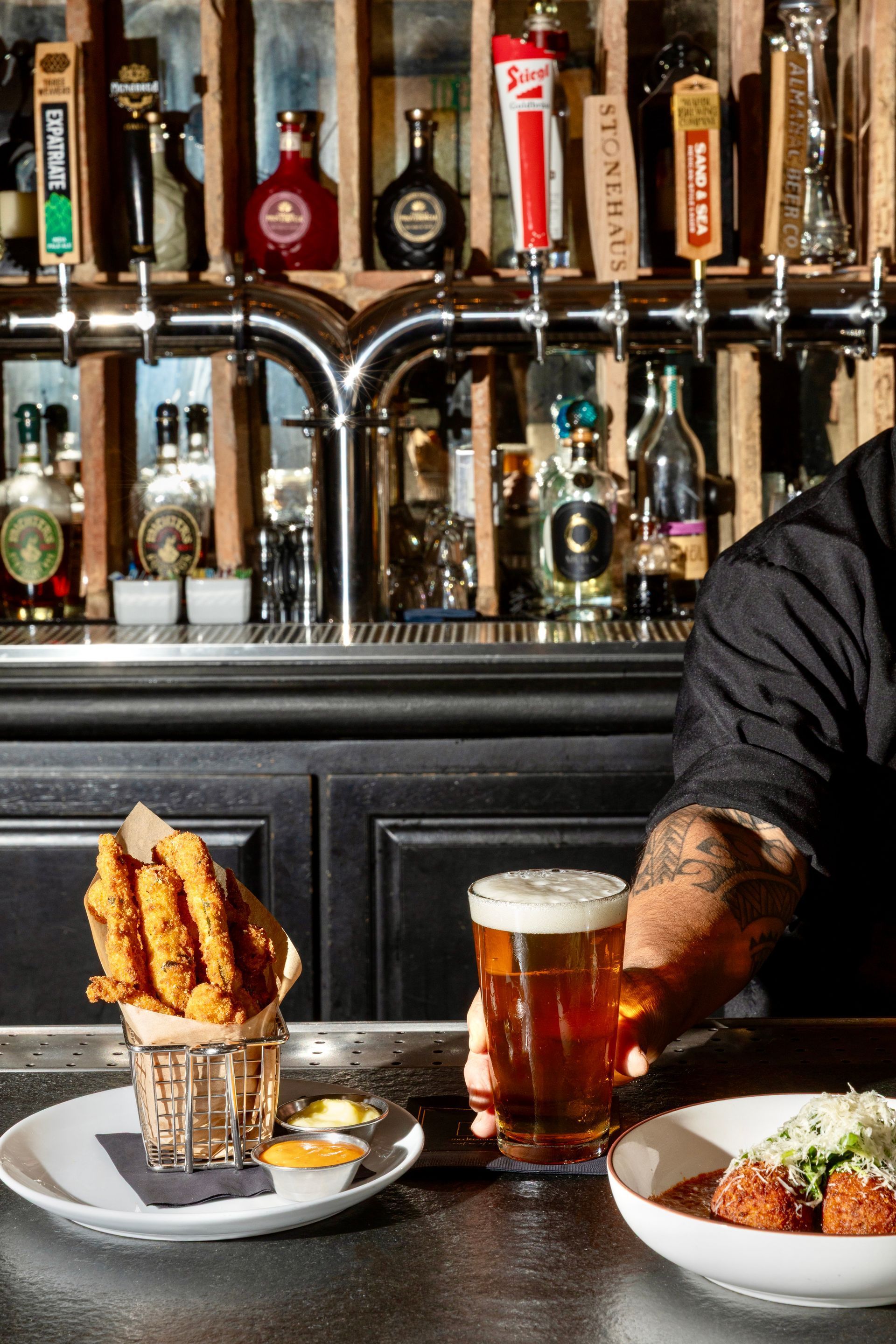 Bar scene with drinks and plated food, including fries and beer in front of a bartender’s arm