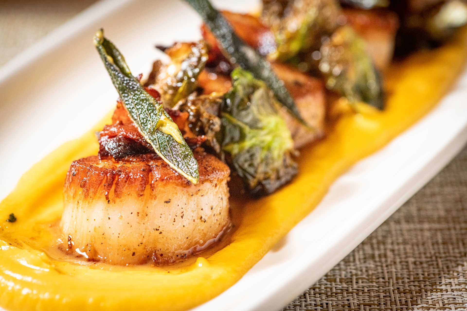 Seared scallops on a bed of yellow sauce, topped with crispy greens and vegetables, served on a white rectangular plate.