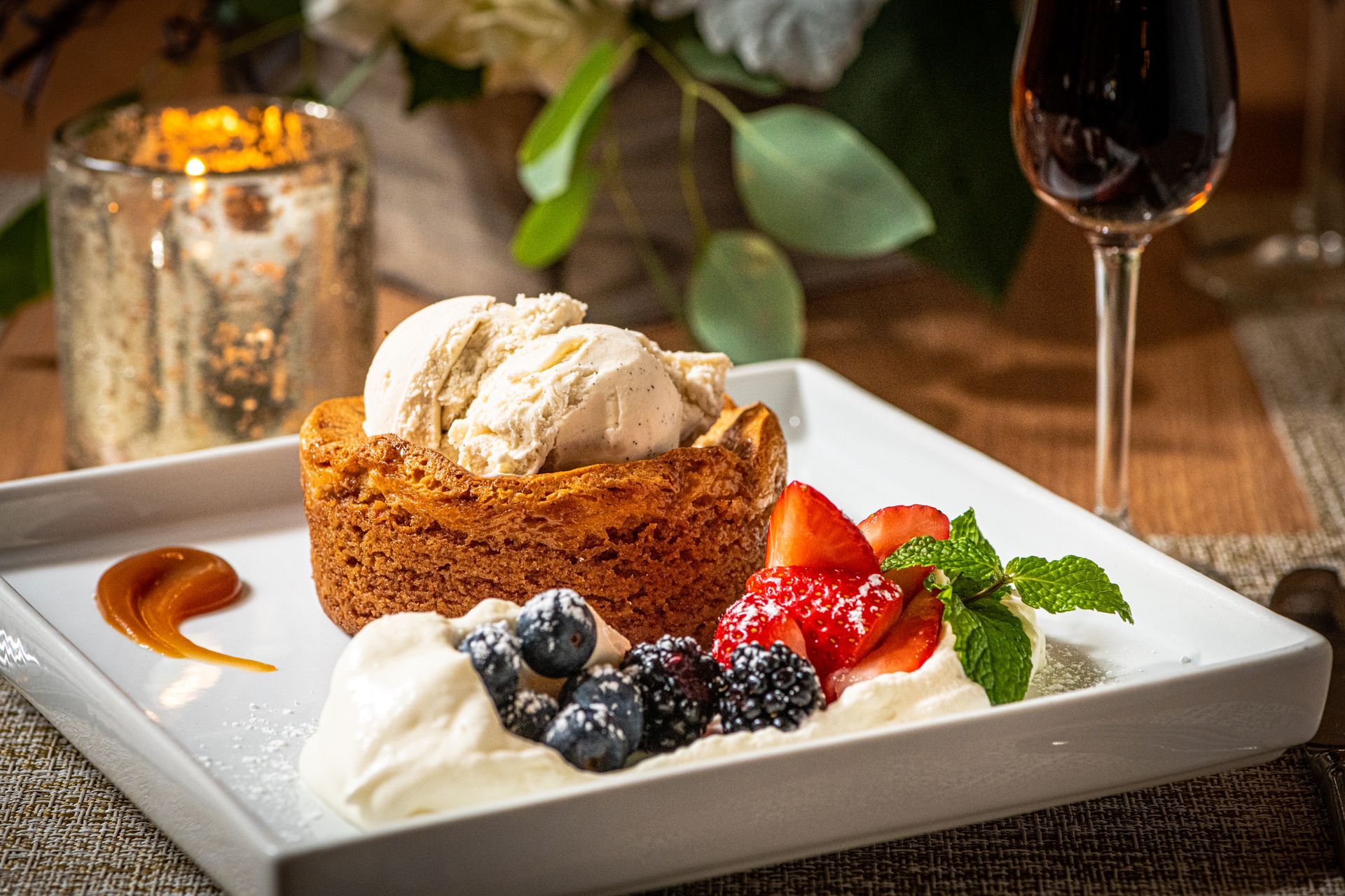 A dessert on a white plate: a cookie with ice cream, berries, whipped cream, and caramel drizzle. A lit candle and wine glass are in the background.