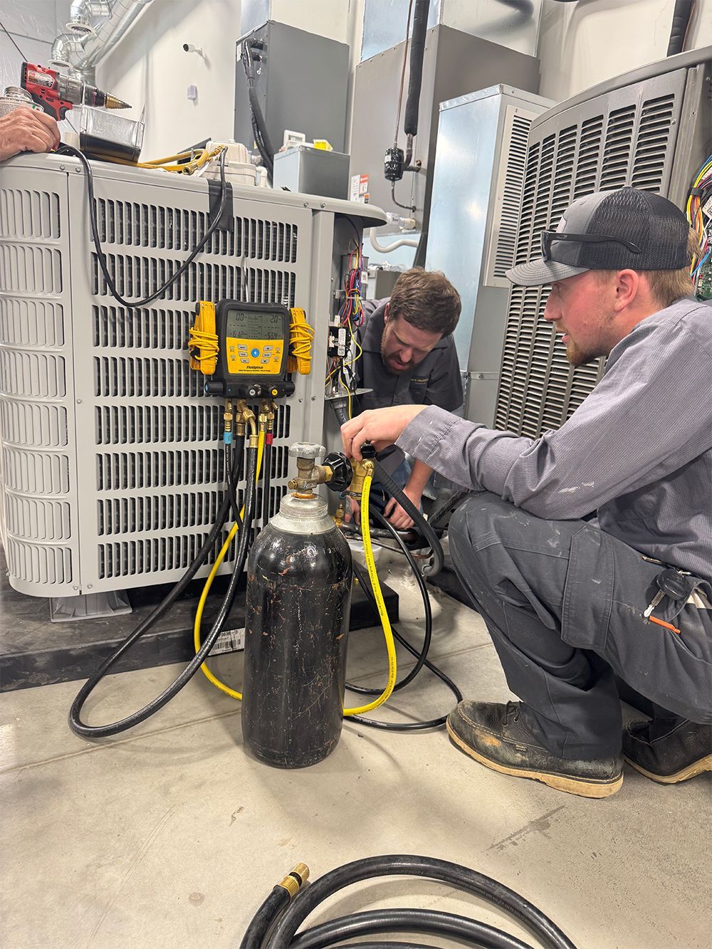 Two HVAC technicians working on an air conditioning unit; one connects gauges, another works on internal components.