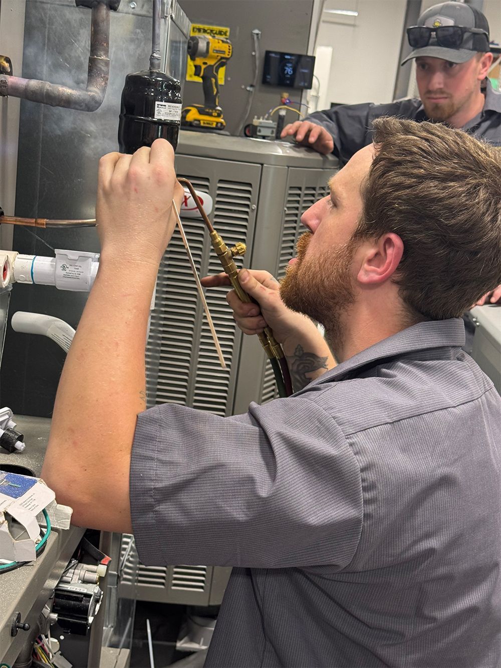 Two HVAC technicians working on a furnace. One uses a tool, other observes.